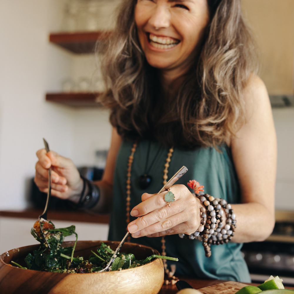Eine lächelnde Frau mittleren Alters steht in einer Küche und richtet einen Salat an. 